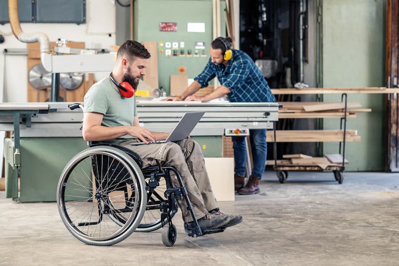 Ein Mann in einem Rollstuhl sitzt mit einem Laptop in einer Werkstatt. Im Hintergrund arbeitet ein Kollege an einer Werkbank.